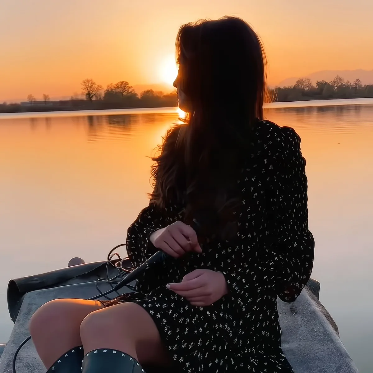 A woman in a floral dress sits on a boat, holding a paddle, gazing at a serene sunset over a calm lake.