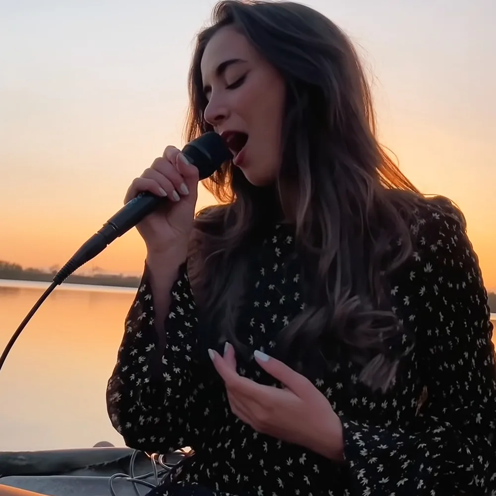 A woman sings passionately into a microphone, her hand on her chest, with a sunset and water in the background.