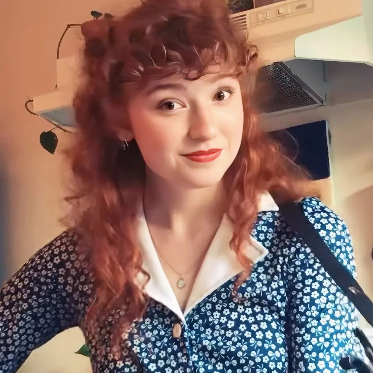 A woman with curly red hair and a blue floral shirt is smiling in a kitchen setting.