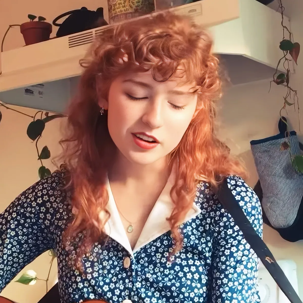 A woman with curly red hair, wearing a blue floral dress, stands indoors with plants and a bag in the background.