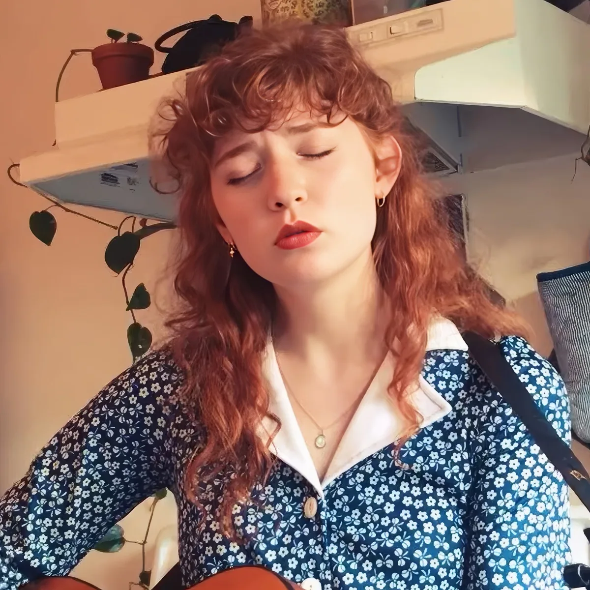 A woman with curly red hair, wearing a blue floral shirt, is playing a guitar with her eyes closed.