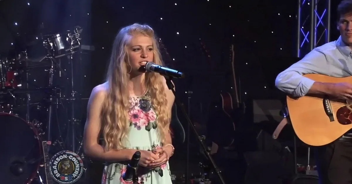 A woman in a floral dress sings on stage with a band, including a guitarist and a violinist.