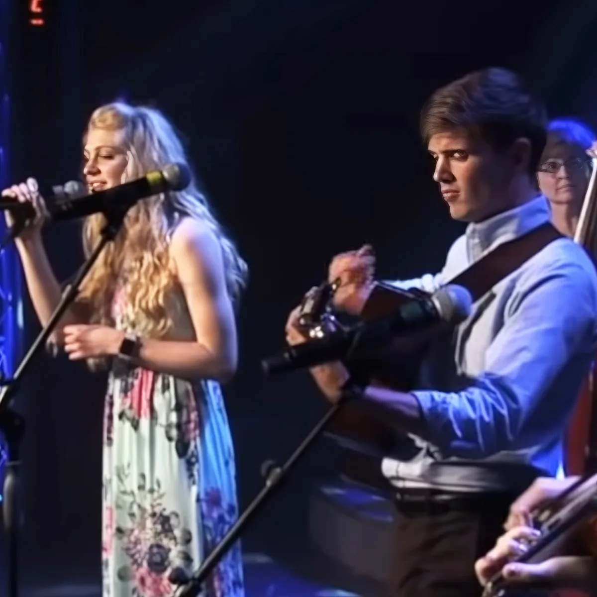 A woman sings into a microphone while a man plays guitar, both on stage with a dark background.