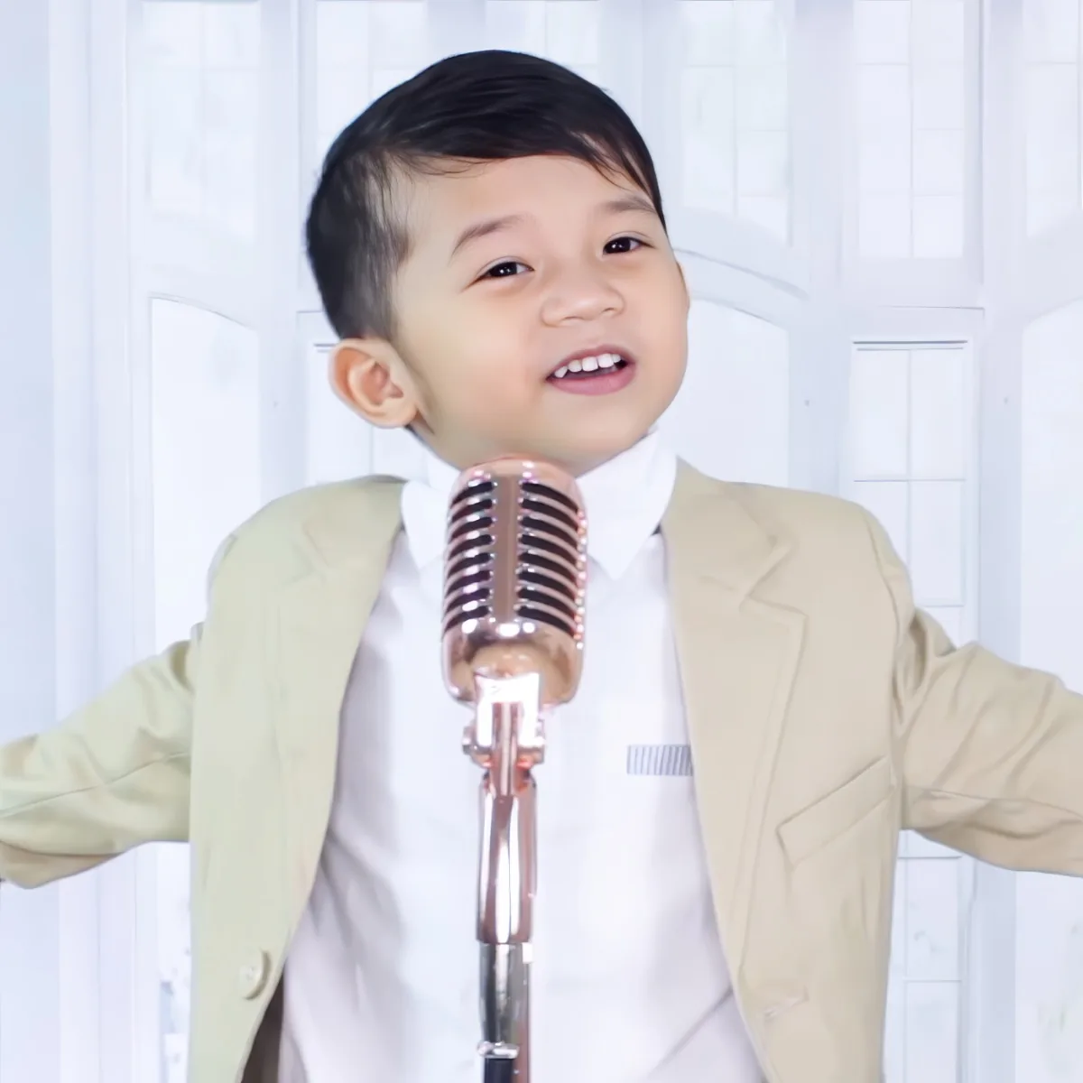 A young boy in a beige suit smiles while standing in front of a vintage microphone.