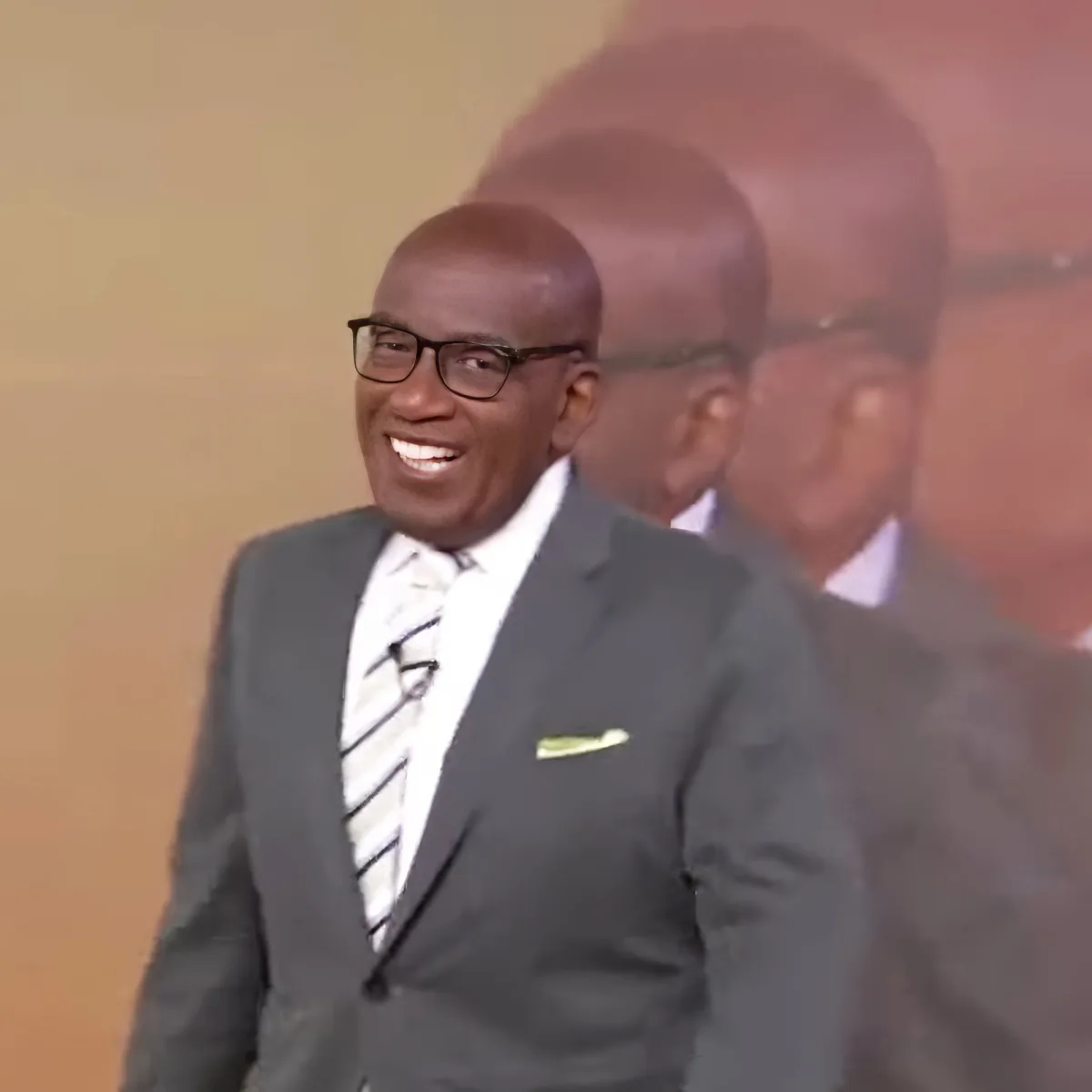 A man in a suit and tie smiles widely, wearing glasses, with a blurred background.