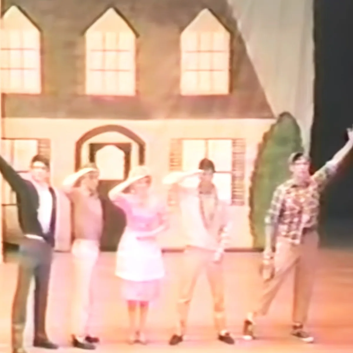 A group of people in vintage attire dances on stage with a house backdrop.
