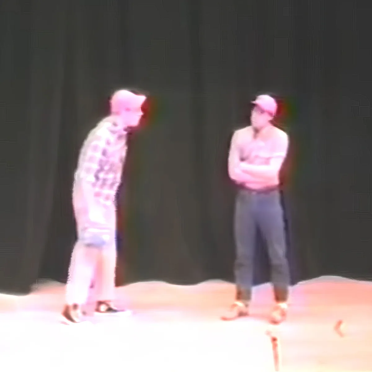 Two individuals stand on a stage, one in a patterned outfit, the other in casual attire, both facing each other.