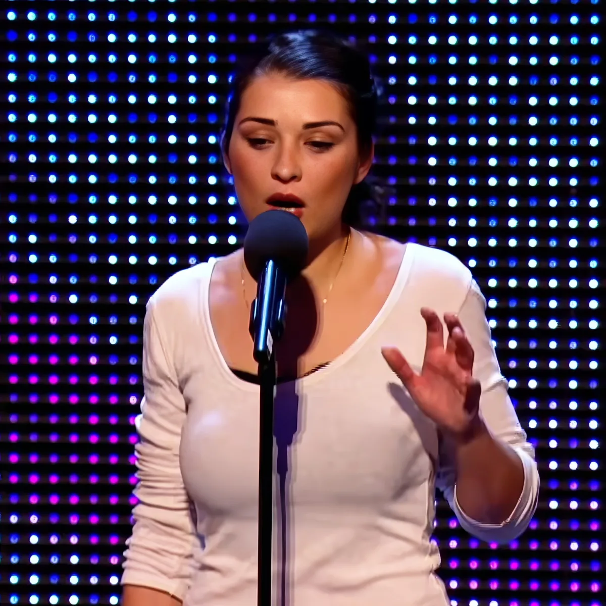 A woman in a white top sings into a microphone, gesturing with her hand, against a backdrop of blue and purple lights.