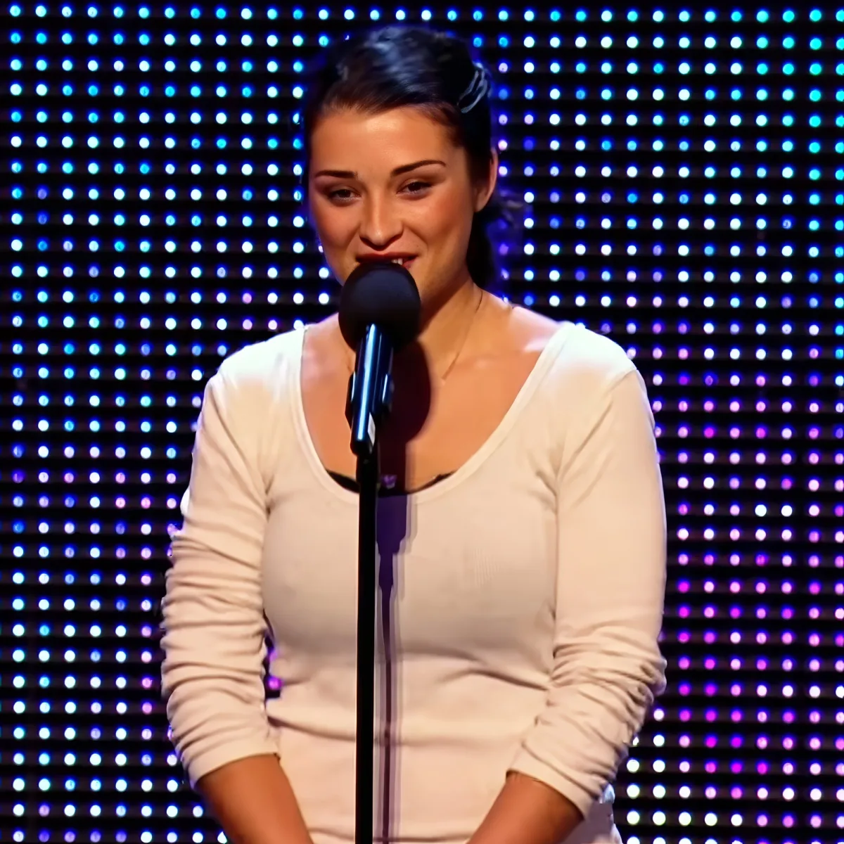 A woman in a white top stands at a microphone in front of a colorful LED backdrop.