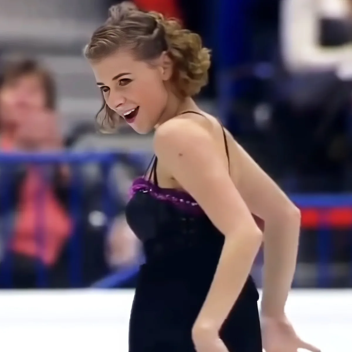 A figure skater in a black dress with purple accents smiles on the ice.
