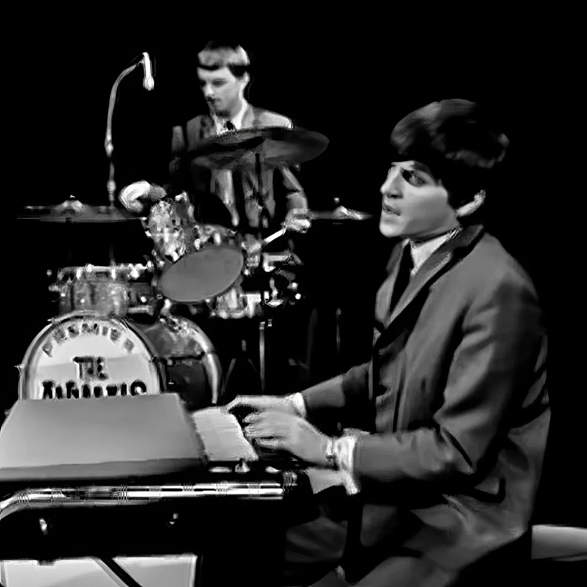 A man in a suit plays a keyboard while another man in a suit plays drums behind him.