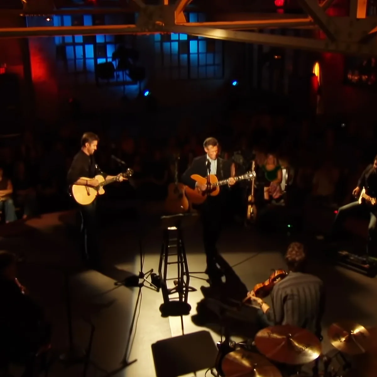 Musicians playing guitars on stage with a drummer and audience in a dimly lit venue.