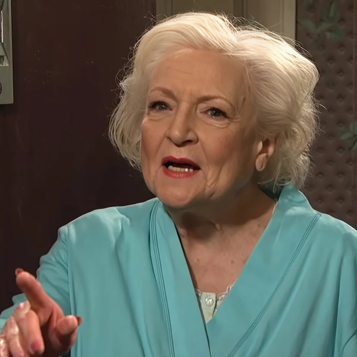 An elderly woman with white hair, wearing a teal blouse, is speaking with her hands gesturing.