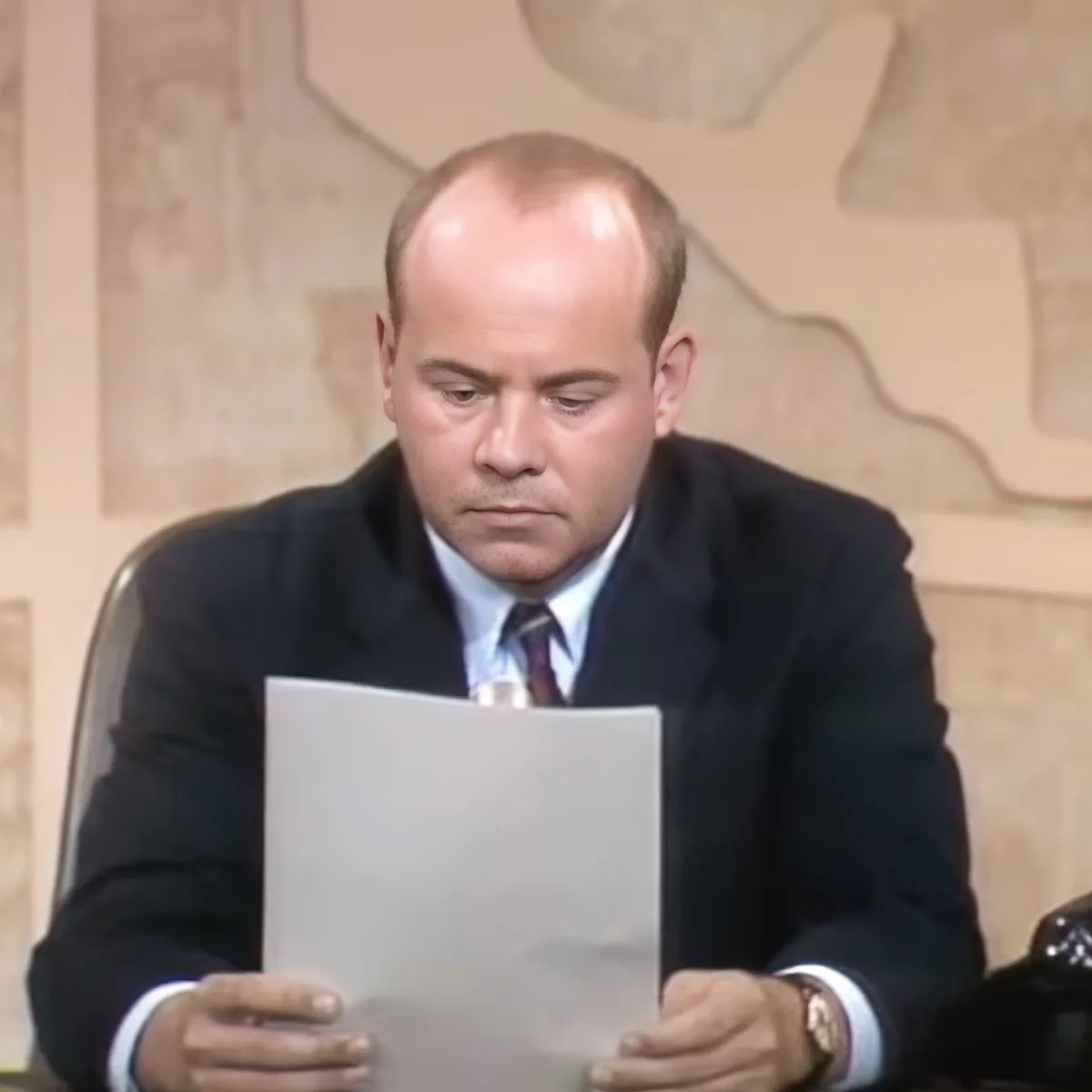 A man in a suit sits at a desk, intently reading a document.