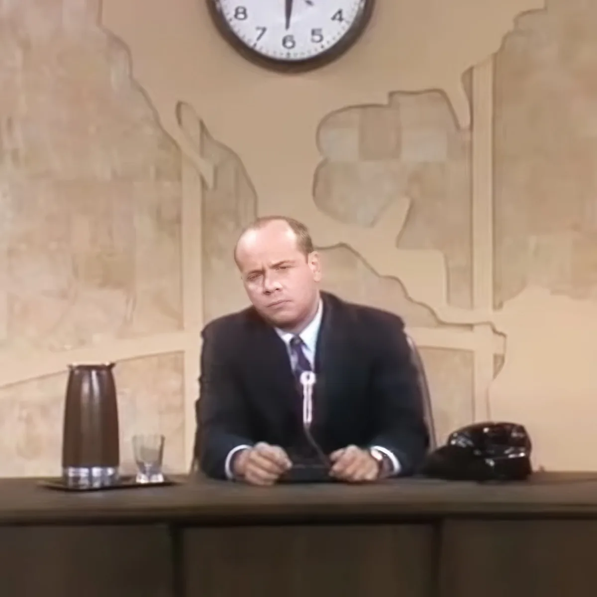A man in a suit sits at a desk with a clock above, appearing to be in a formal setting.