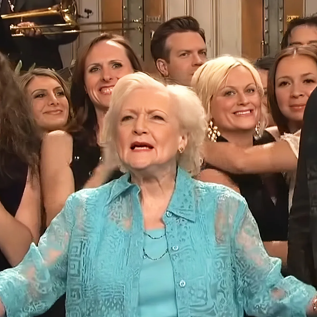 An older woman in a blue shirt sits in front of a group of people, seemingly reacting to something.