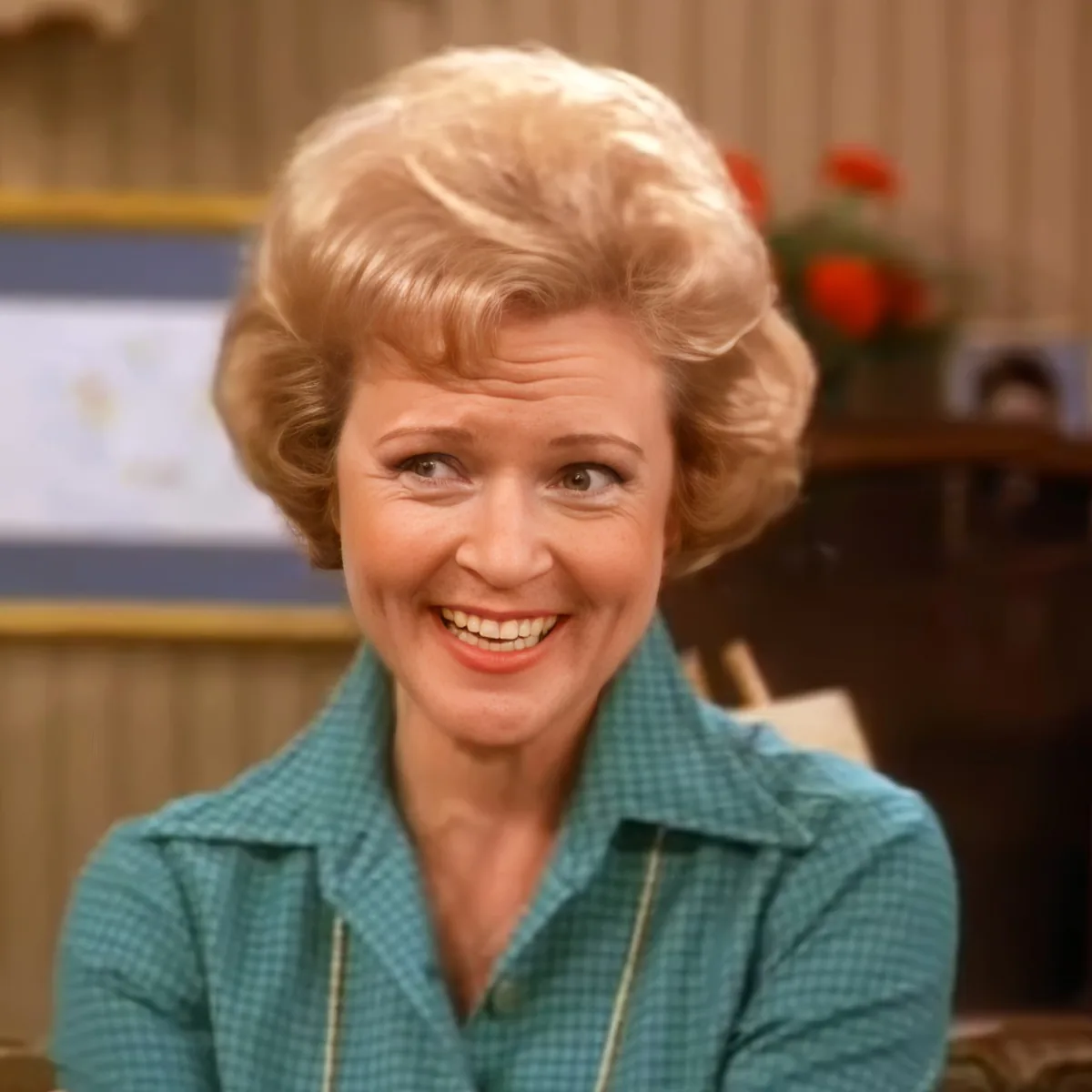 A smiling woman with blonde hair in a teal checkered shirt, sitting in a room with a map and fruit in the background.