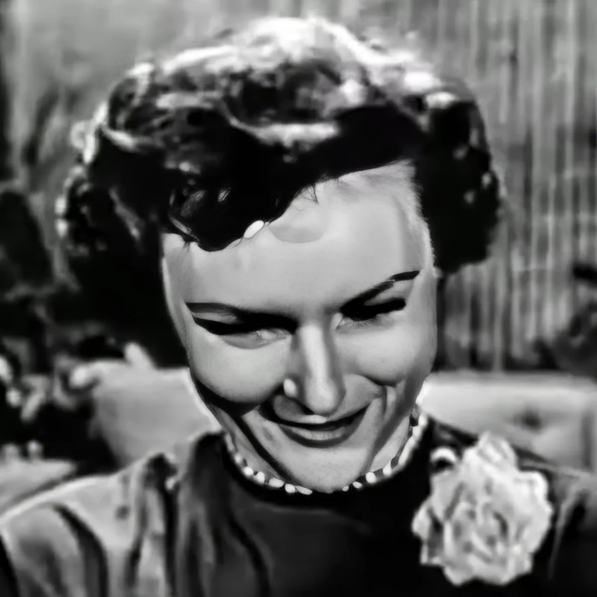 A woman with a pearl necklace and a flower in her hair is smiling.