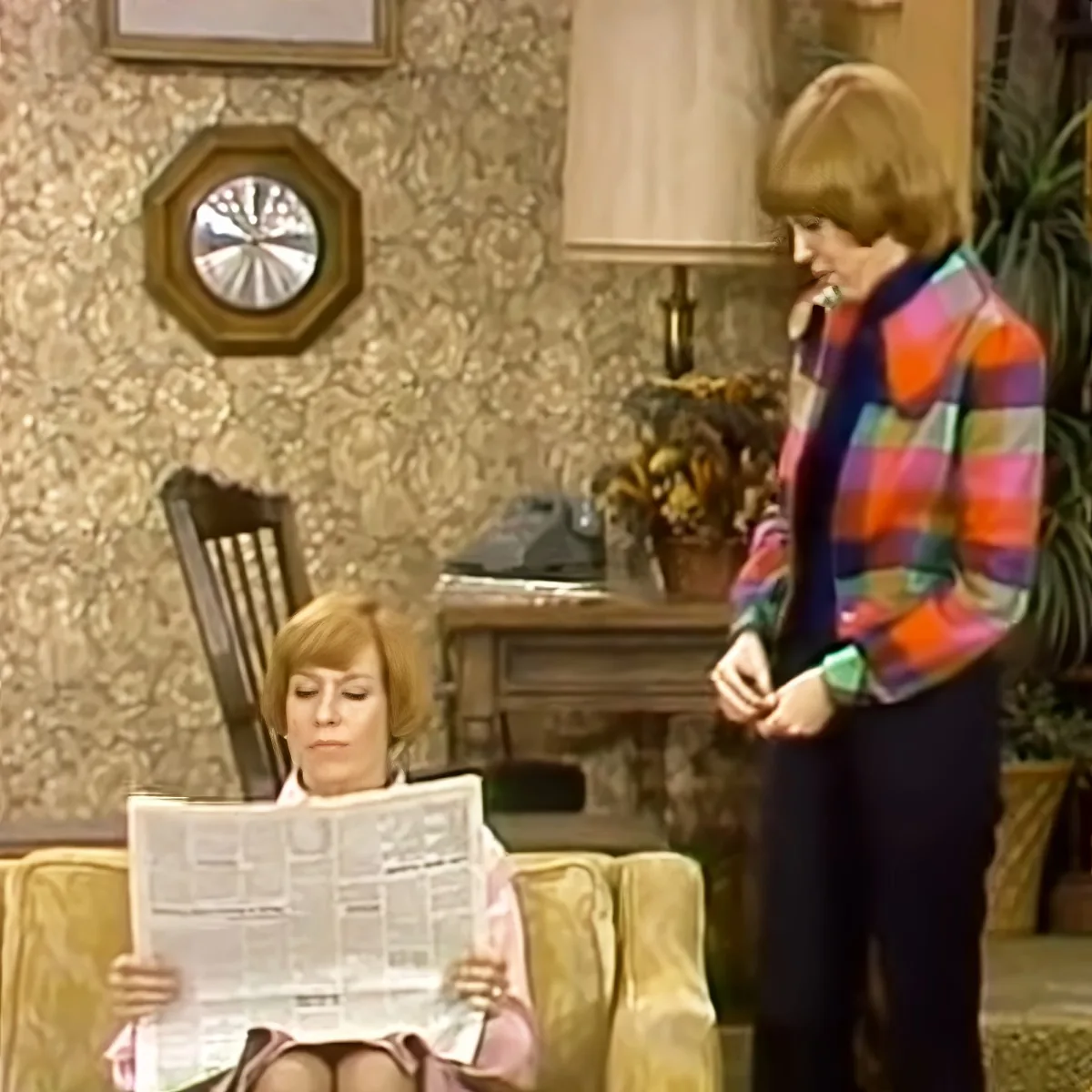 Woman in colorful plaid jacket stands, while another reads newspaper in a cozy living room setting.