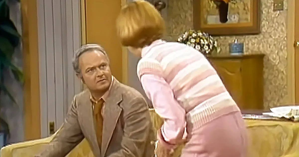 A woman in a pink striped sweater leans over a man in a suit, who looks surprised, in a warmly lit room.
