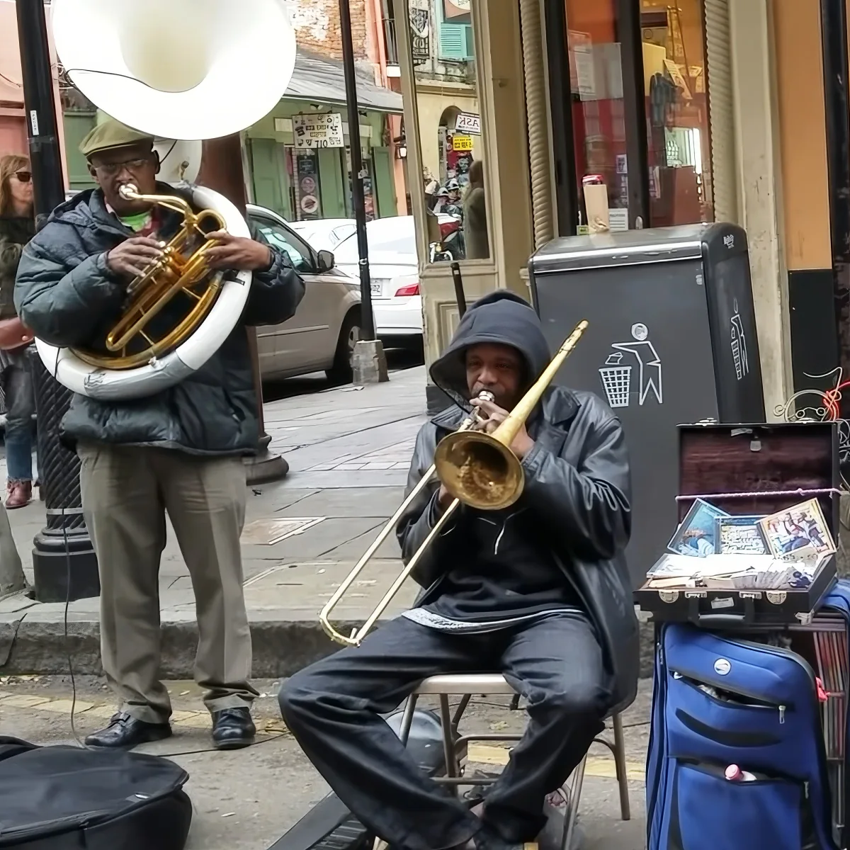 A man in a hoodie plays a trombone on a street, with another musician playing a sousaphone nearby.