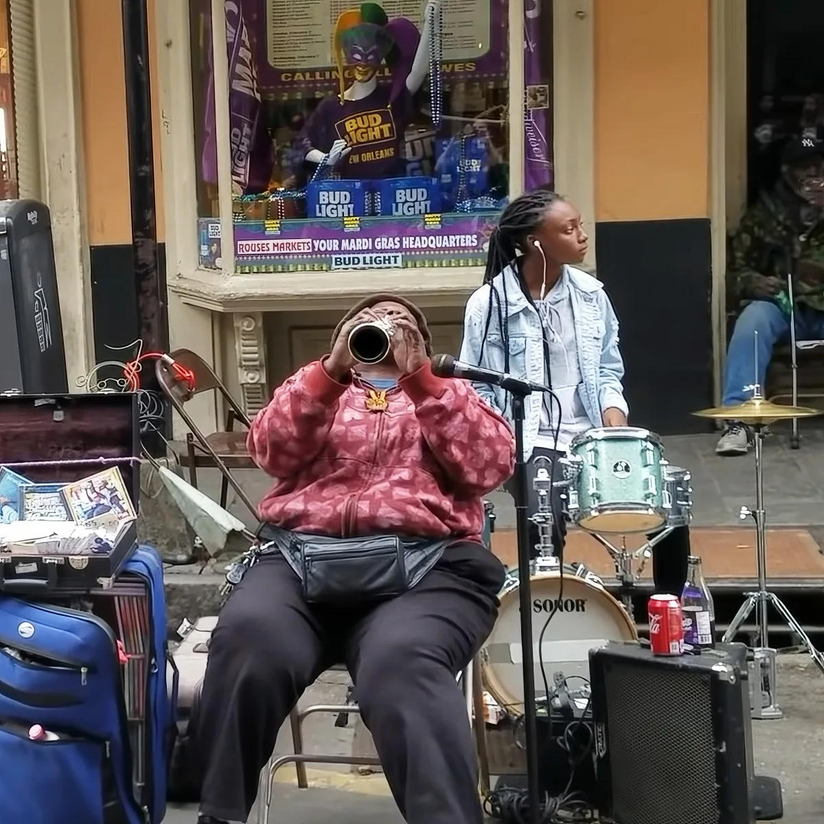 A person in a red jacket plays a trumpet while a drummer sits behind them, with a Bud Light advertisement in the background.