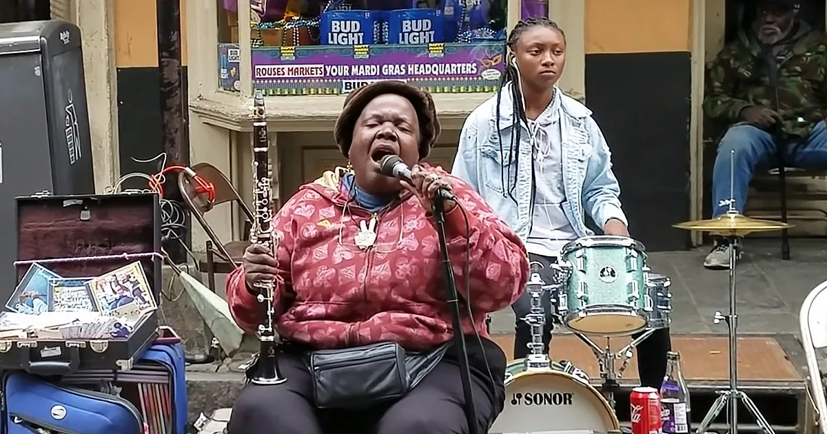 A woman plays clarinet while singing into a microphone, accompanied by a drummer.