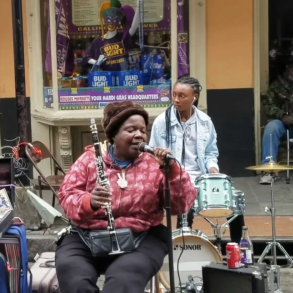 A woman plays clarinet and sings into a microphone while a drummer stands behind her.