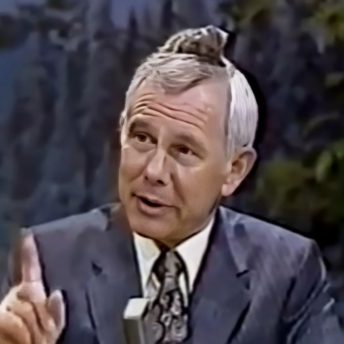 A man in a suit and tie is speaking, gesturing with his right hand.