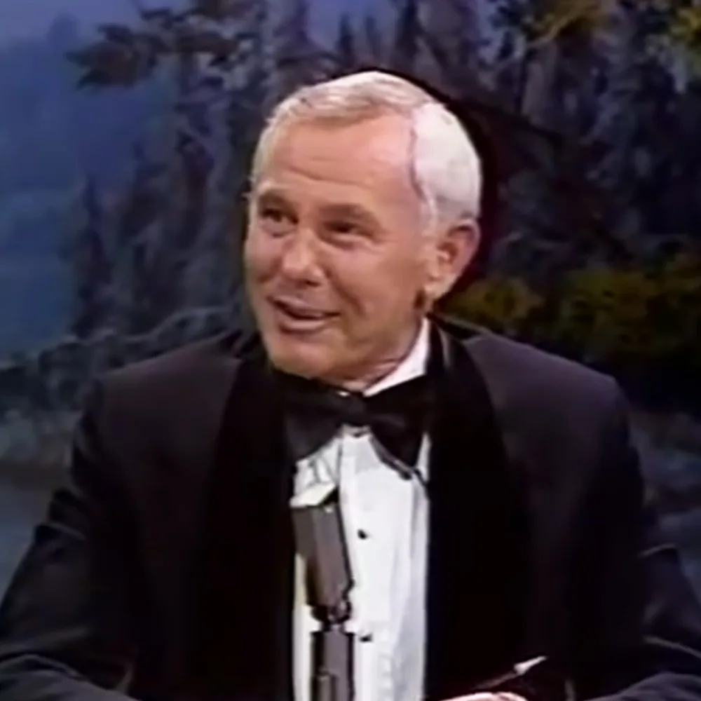 A man in a tuxedo smiles while seated, with a scenic backdrop behind him.
