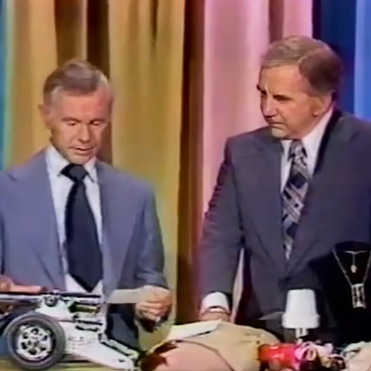Two men in suits stand at a table with a model motorcycle and jewelry, possibly on a game show set.