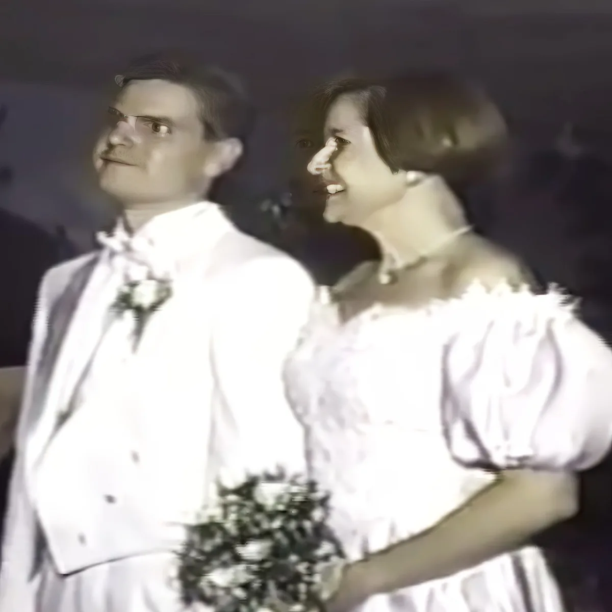 A couple in formal attire, the woman in a white dress holding a bouquet, the man in a white suit with a boutonniere.
