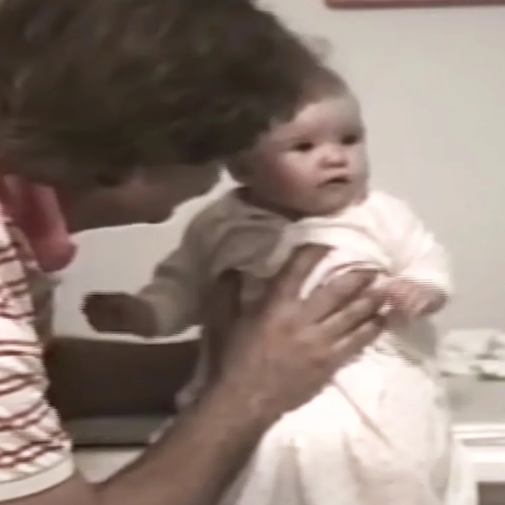 A person holds a baby in a white outfit, with a blurred background and a chair visible.