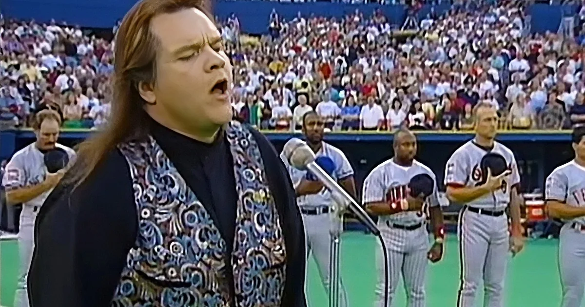 A man in a patterned vest sings into a microphone at a baseball game with players and a crowd in the background.
