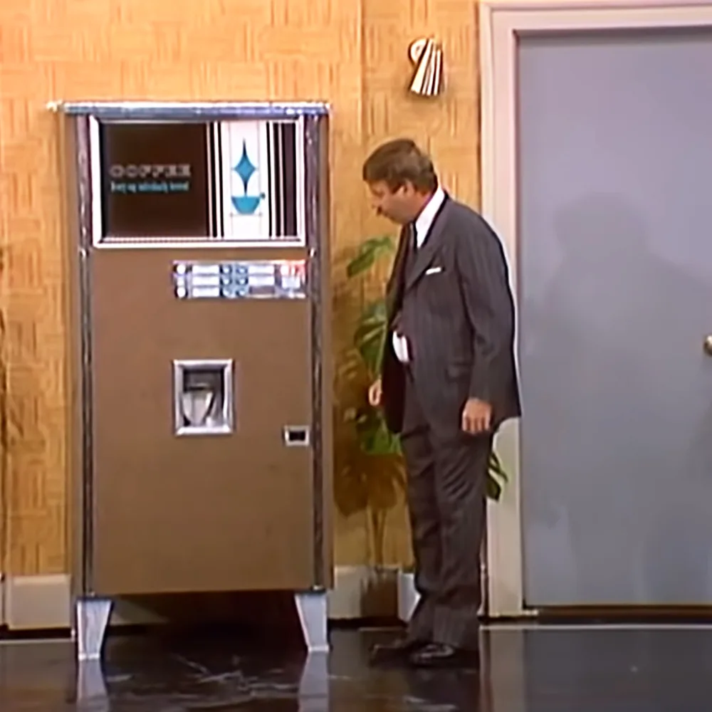 A man in a suit stands beside a vintage coffee vending machine in a room with wood paneling.