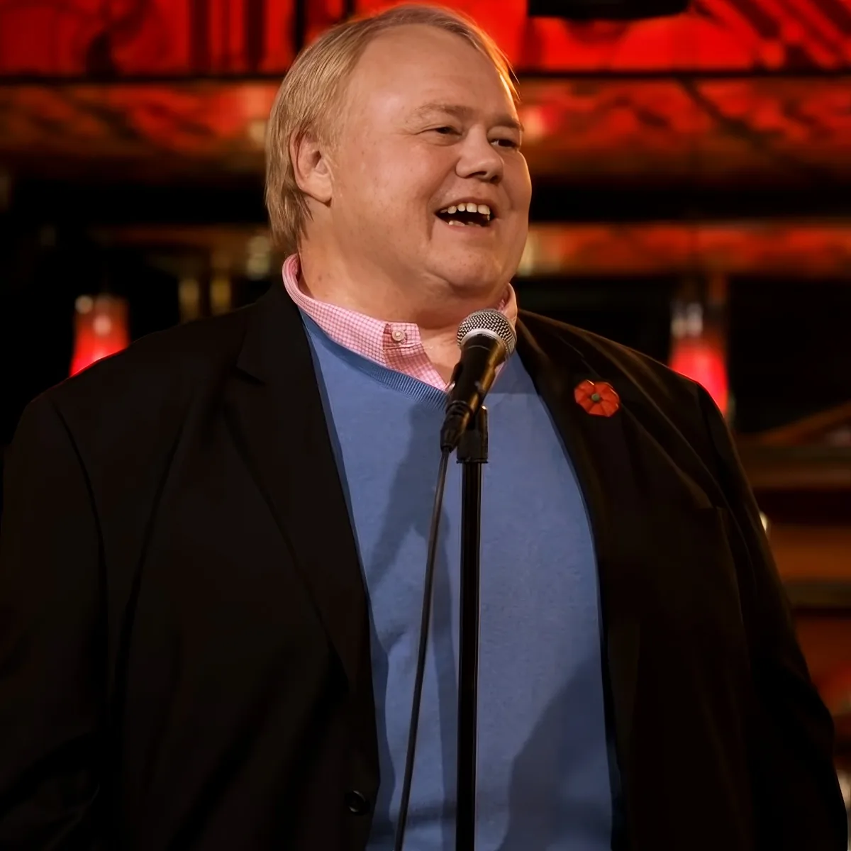 A man in a black blazer and blue shirt stands at a microphone, smiling, with a red poppy on his lapel.