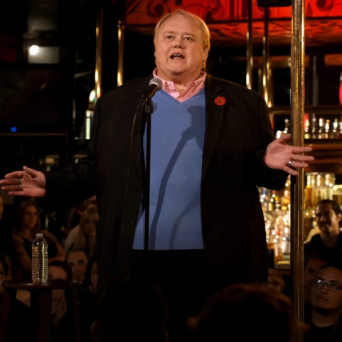 A man in a suit and sweater stands on stage with a microphone, gesturing to an audience in a dimly lit venue.