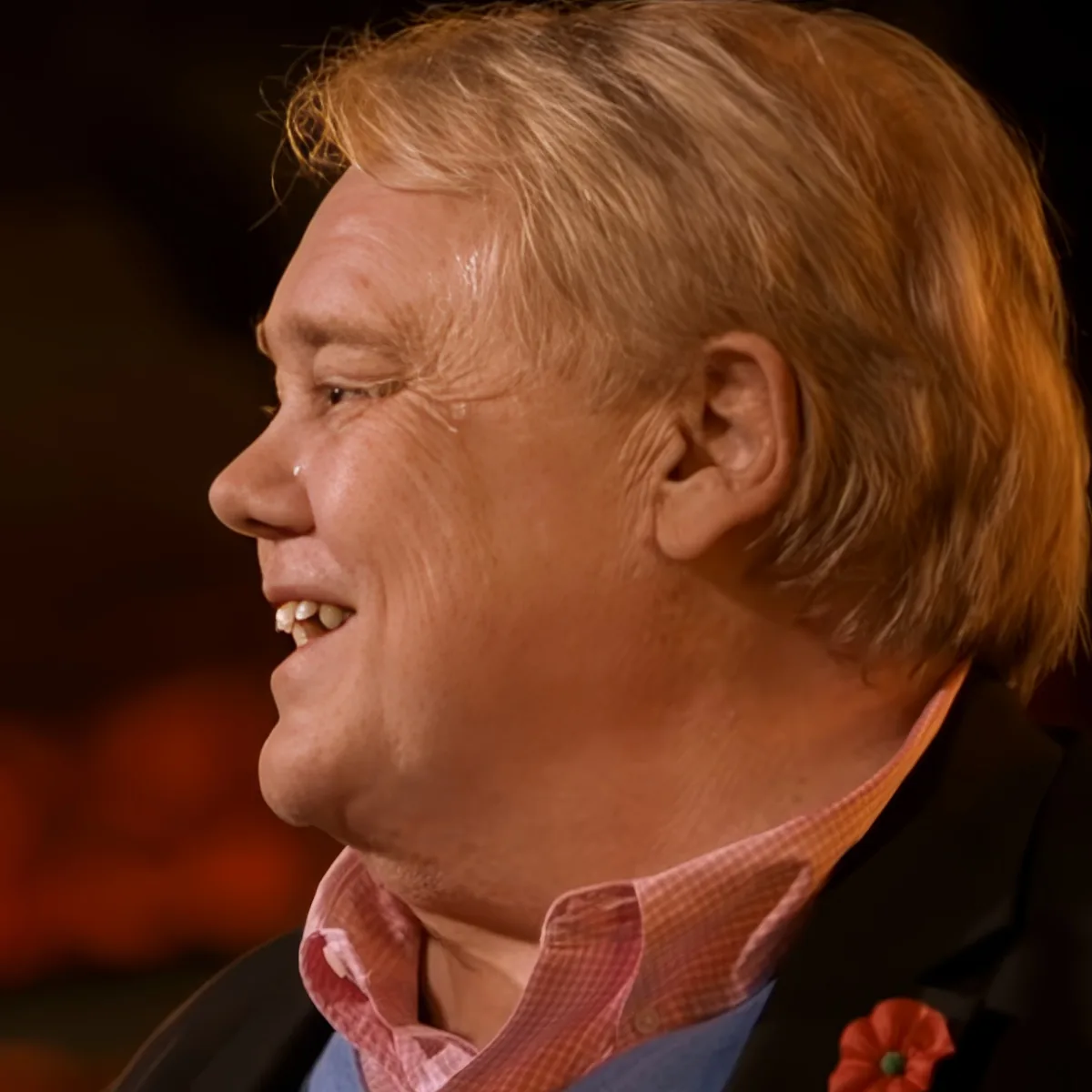 A person with light brown hair, wearing a red checkered shirt and a dark blazer, is smiling and looking to the left.