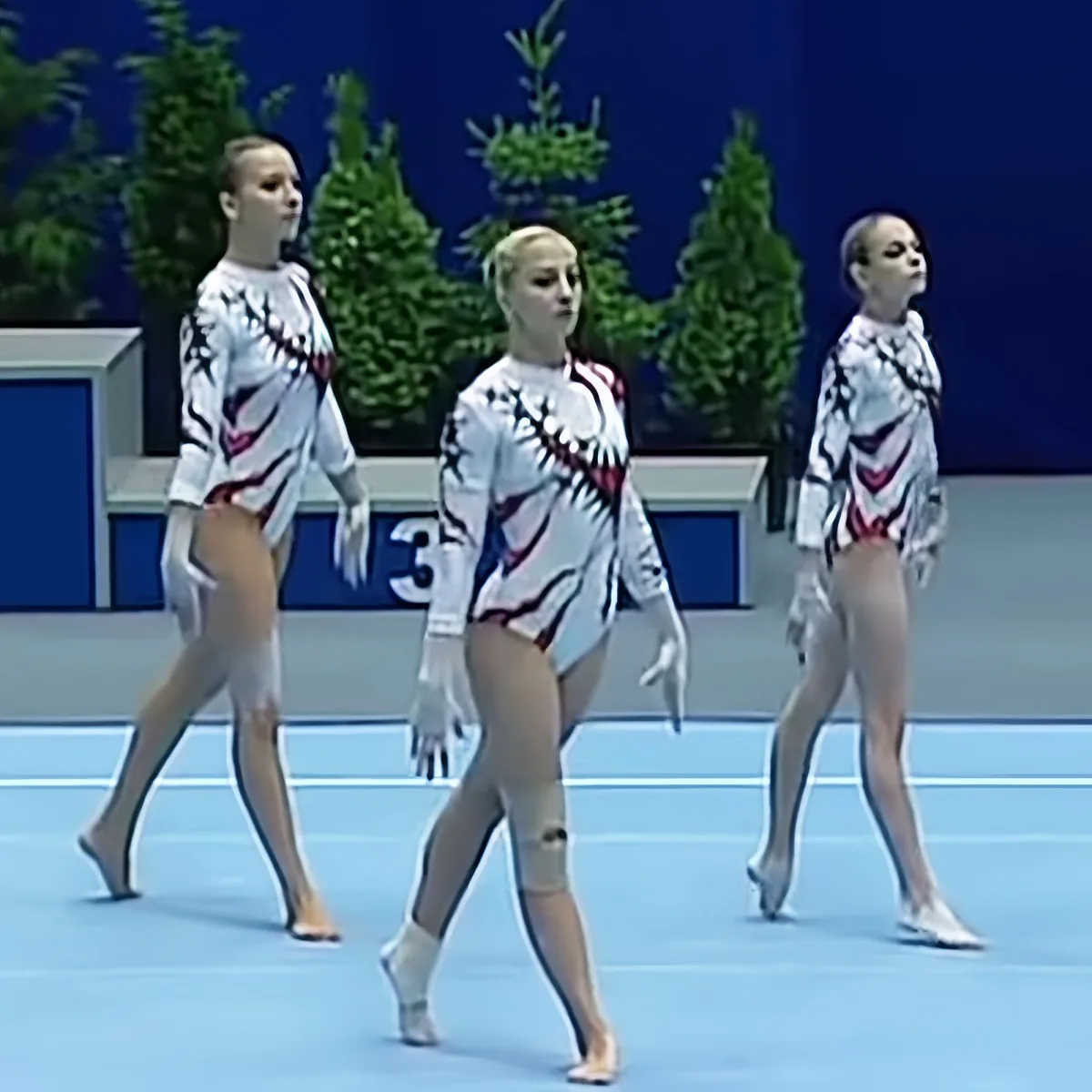 Three synchronized gymnasts in matching white and red leotards perform a routine on a blue floor.