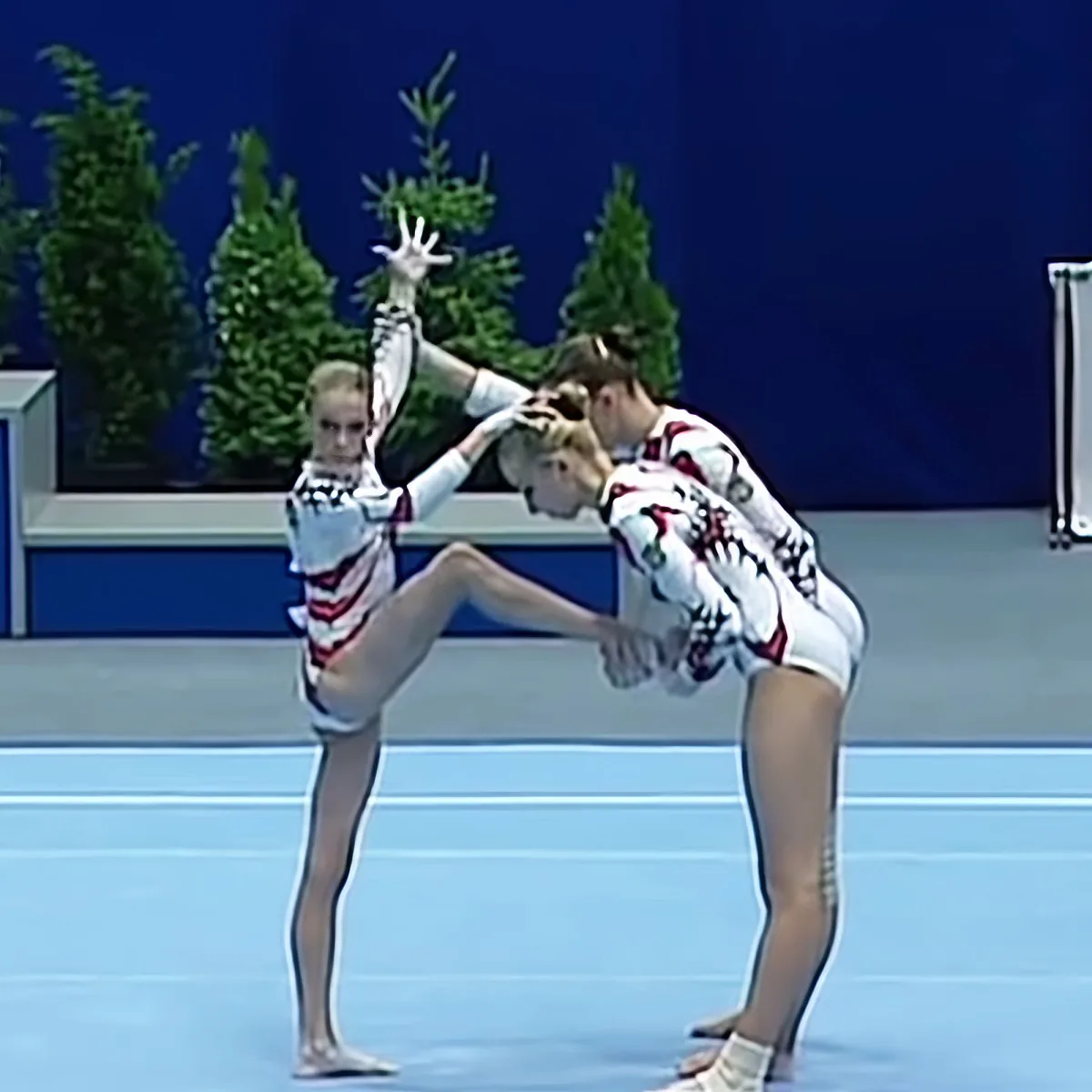 Two gymnasts in synchronized pose, one supporting the other in a split, on a blue mat.