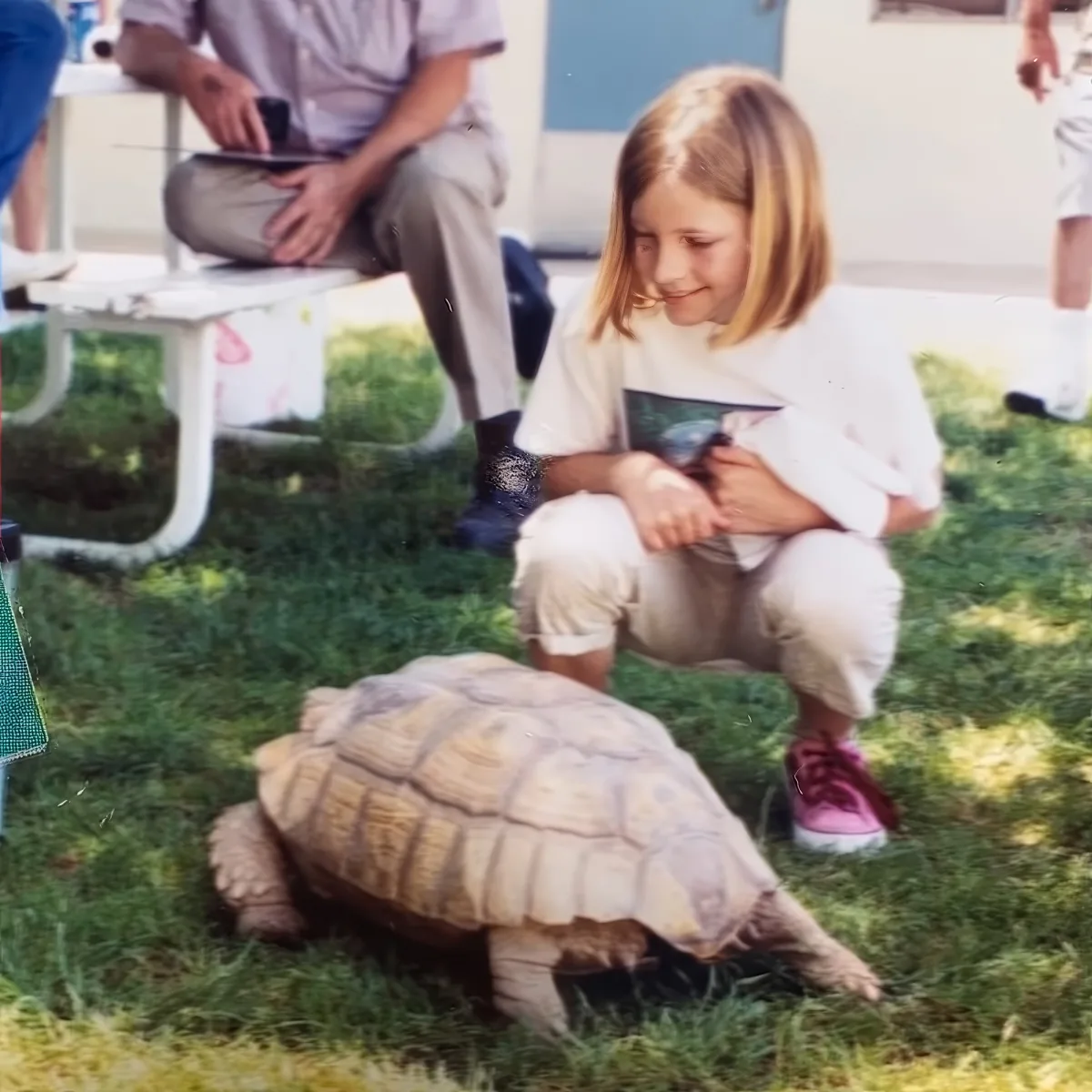 A young girl crouches near a large tortoise on grass, smiling, with a man sitting on a bench in the background.