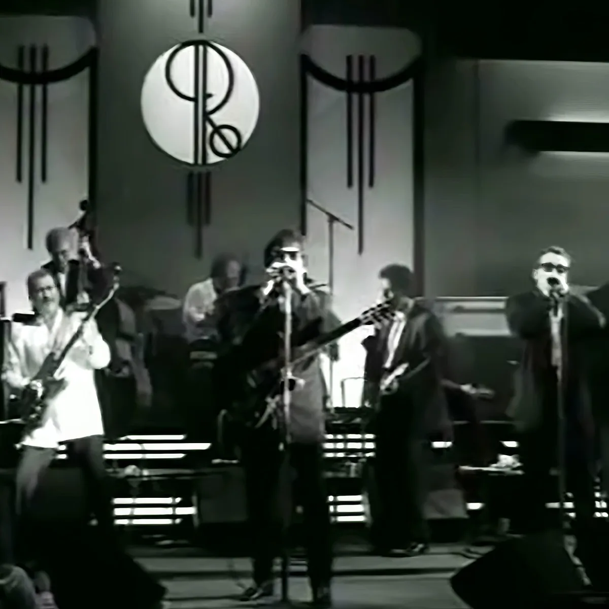 A band performs on stage with microphones, guitars, and a large circular logo in the background.