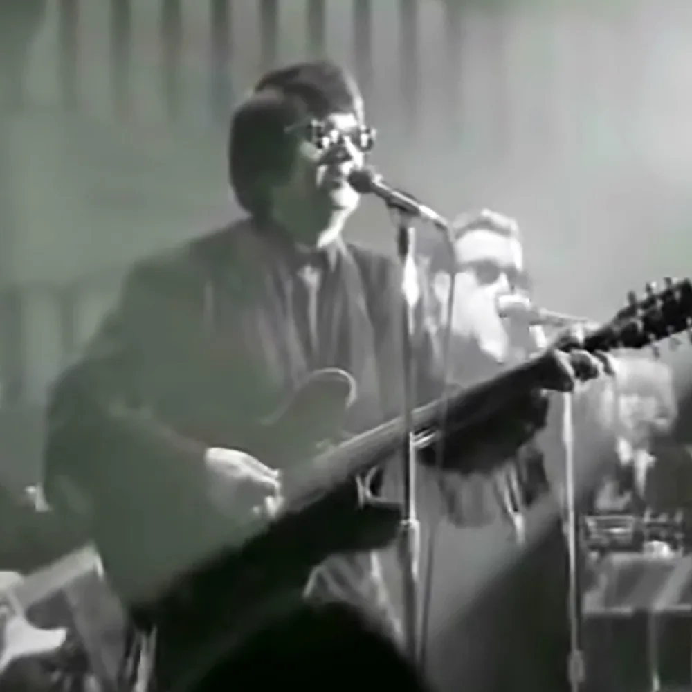 A man in glasses plays guitar and sings into a microphone on stage.