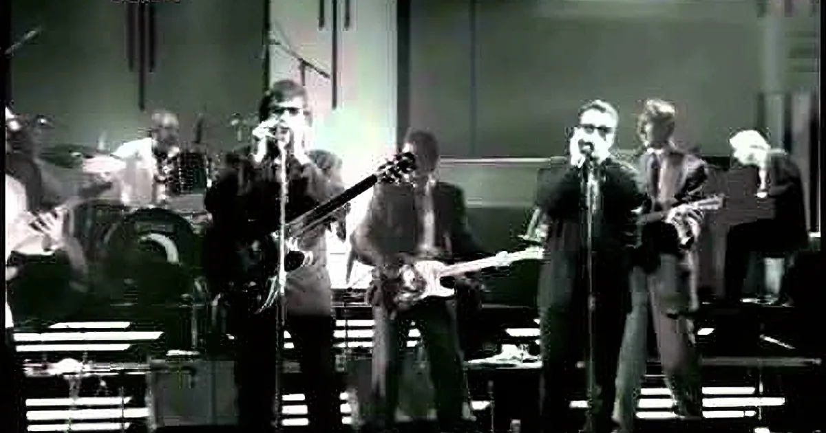 A person wearing sunglasses sings into a microphone while holding a guitar, with a blurred background.