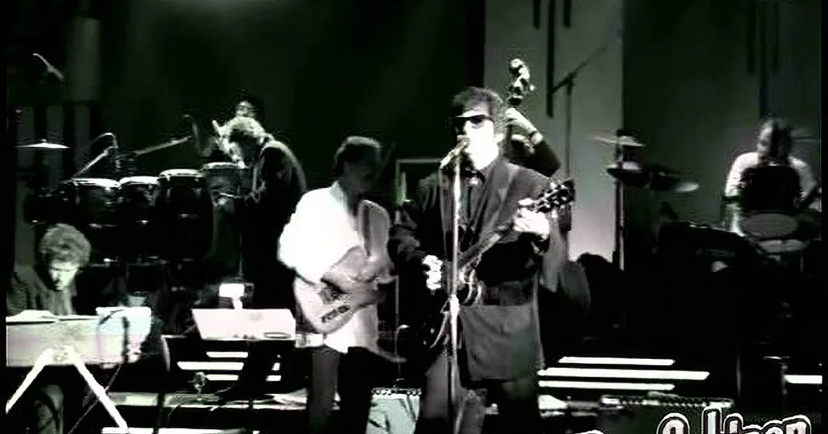 A man in a suit and sunglasses sings into a microphone, holding a guitar.