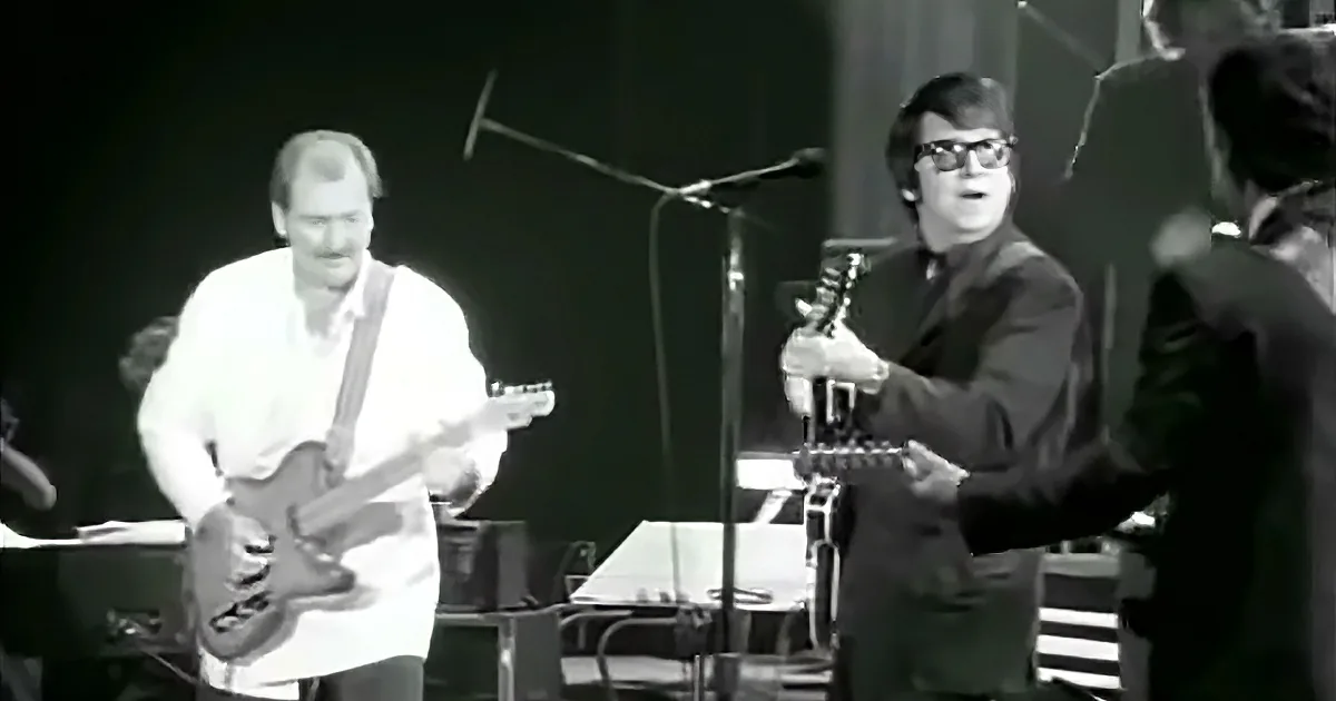 A man in a white shirt plays guitar while another in a dark suit sings into a microphone.