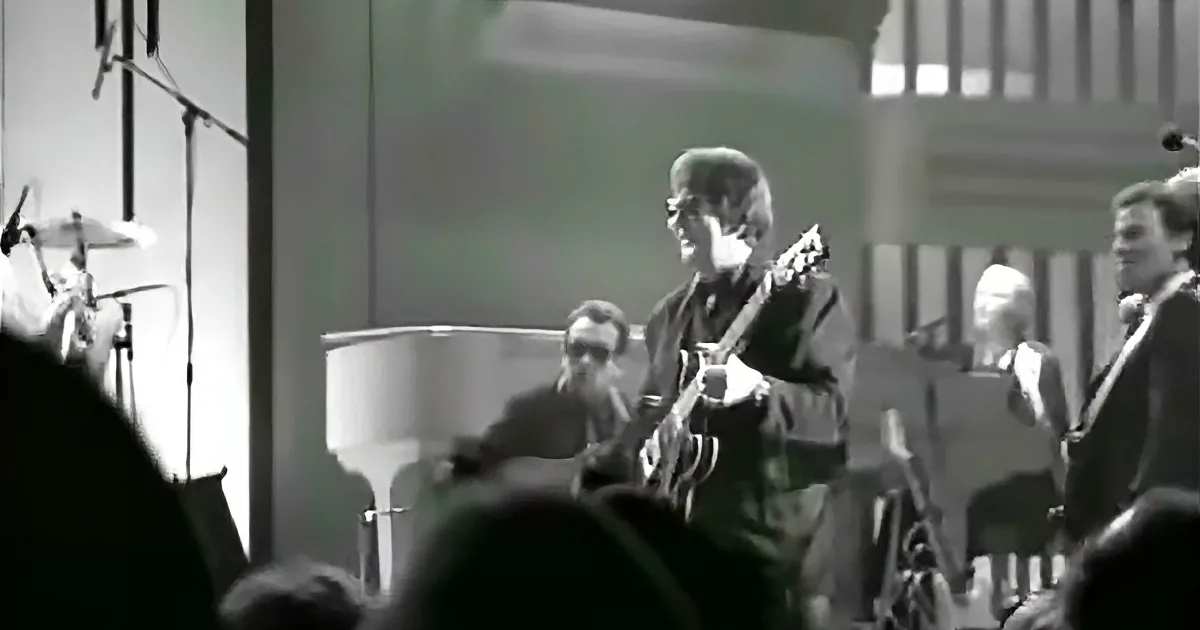 A musician plays guitar on stage, surrounded by other musicians and equipment.
