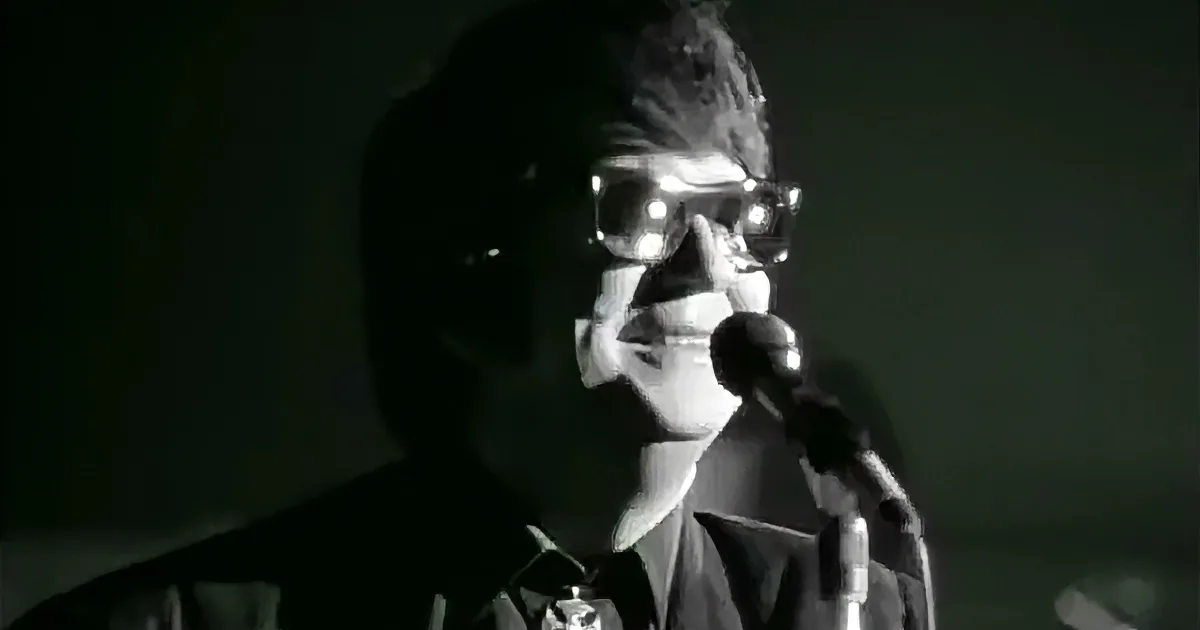 A person wearing glasses and a suit is singing into a microphone, with a dark background.