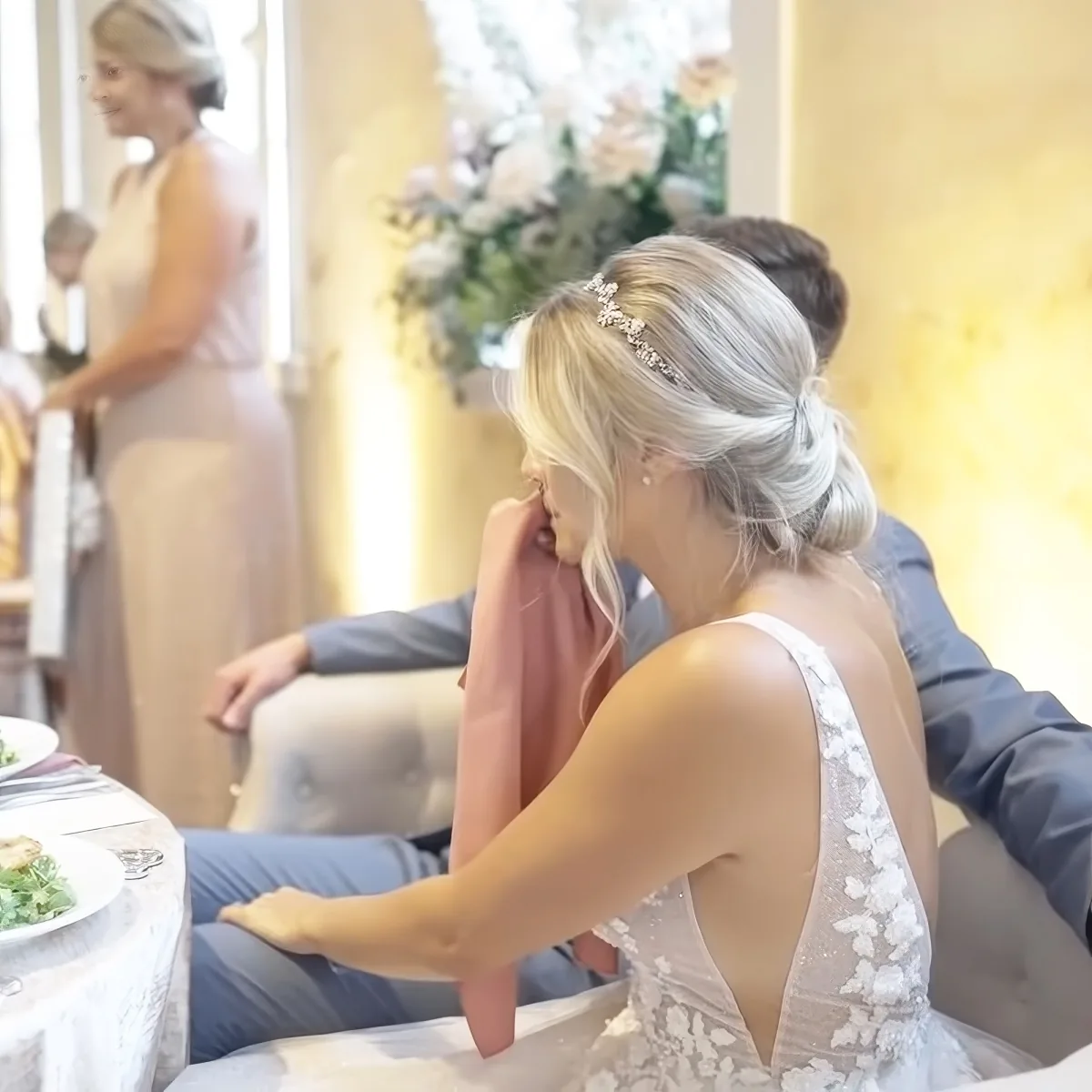 A bride in a white dress with a lace back sits at a table, serving food to a seated couple.