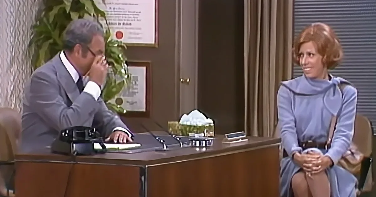 A man in a suit sits at a desk, appearing to think, while a woman in a blue dress sits across from him, smiling.